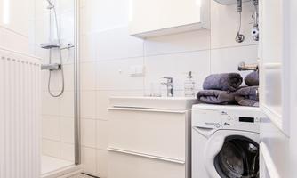 A modern bathroom with a shower and a sink. A washing machine stands next to a stack of towels.