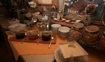 A breakfast buffet with various jams, cereals, and fresh rolls. Plates and bowls are also provided on a table.