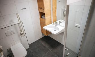 A modern bathroom with a shower, a sink, and wooden shelves. The walls are light tiled and the floor is dark.