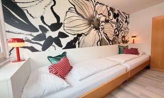 A modern bedroom with two beds and colorful pillows. The wall is decorated with a striking floral pattern.