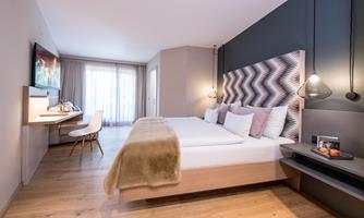A modern bedroom with a large bed and minimalist design. There is a desk, a TV, and plenty of daylight from the windows.