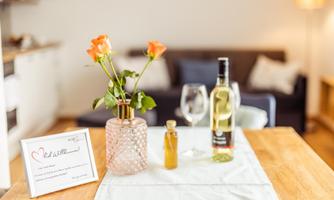 A rustic table with a vase full of roses, a bottle of wine, and glasses. In the background, a cozy seating area is visible.