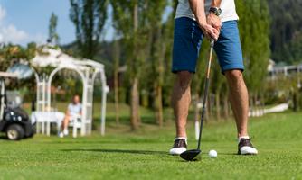 A golfer is standing on the grass and preparing to hit the ball. In the background, there is a relaxed atmosphere with a table and chairs visible.