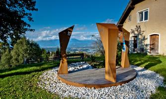 A modern metal sculpture stands on a wooden platform surrounded by gravel. In the background, gentle hills and a blue sky are visible.