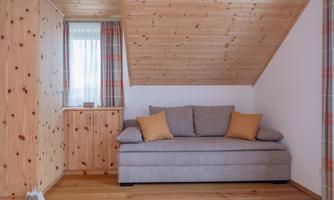 A cozy living room with a gray sofa and orange cushions. The walls are made of wood, and there is a window with a sheer curtain.