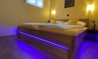 A cozy bedroom with a wooden bed and yellow walls. The atmosphere is enhanced by the purple LED lighting under the bed and the soft wall lighting.