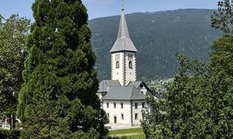 A picturesque landscape with a tall, slender tree and a church in the background. The church has a distinctive tower and is surrounded by green grass and trees.