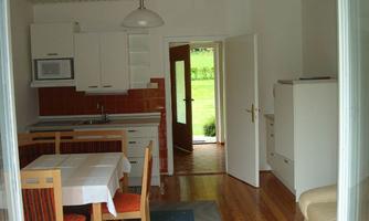 A cozy kitchen area with hardwood flooring and a table for four people. There are large windows that provide a view of the green outdoor area.