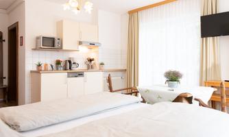 A bright room with two beds and a small kitchenette. A table with chairs and windows with plenty of natural light can also be seen.
