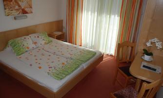 A cozy bedroom with a large bed and colorful pillows. Bright curtains are at the window and there is a small table with a chair.