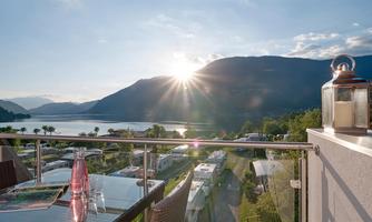A picturesque view from a balcony overlooking a lake and mountains in the background. The sun is just rising, casting warm light over the scene.