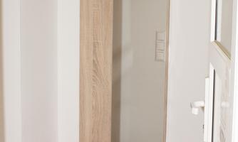 A simple entrance area with a wooden sign, a mirror, and a coat hook. The bench has red cushions and a white drawer underneath.