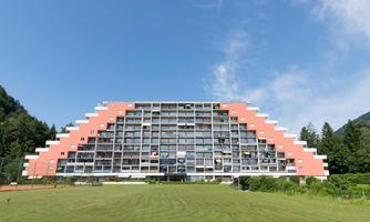 A modern building with striking architecture in the shape of a house. It is surrounded by green meadows and trees, under a clear blue sky.