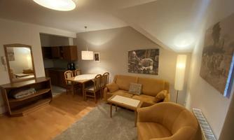 A cozy living room with beige furniture and a dining table. Pictures hang on the wall, and a carpet creates a warm atmosphere.