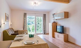 A cozy living room with a dining table and elegant furnishings. Large windows provide a lovely view of the green surroundings.