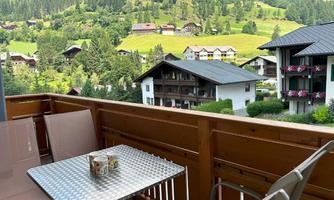 A cozy balcony with a table and two chairs. In the background, green hills and traditional chalets can be seen.