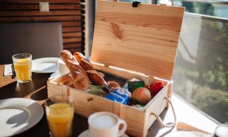 A wooden box with fresh rolls, fruit, and drinks is on a table. Next to it are plates and glasses with orange juice and coffee.