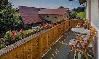 A cozy balcony with a wooden railing and colorful flowers. In the background, rustic buildings and a green landscape can be seen.