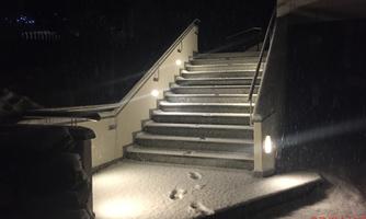 A illuminated staircase leads upwards, surrounded by fresh snow at night. Snowflakes gently fall, creating a calm atmosphere.