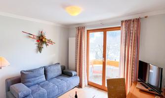 A cozy living room with a blue sofa and a table. Large windows provide plenty of daylight and a view outside.