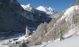 A picturesque winter landscape with snow-covered mountains and a small church in the valley. The trees are covered with snow and the sky is blue and clear.