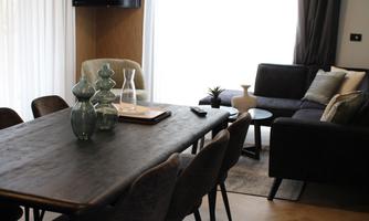 A modern living room with a large dining table and elegant chairs. The rooms are bright and well-lit with a television on the wall.