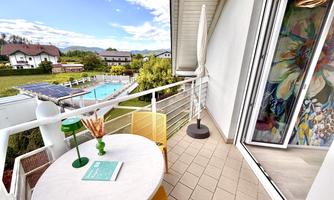 A cozy balcony with a round table and colorful chairs. In the background, a beautiful garden with a pool can be seen.