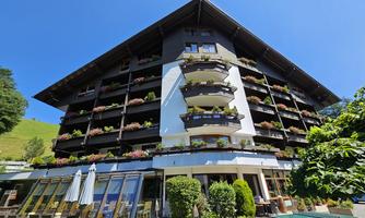 A cozy hotel building with balconies full of flowers. It is located in a green setting under a clear blue sky.