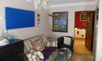 Seating area with sofa, armchair and glass table, dresser with tea station, blue infrared panel and framed wall artwork; open passage to the room’s entrance area.