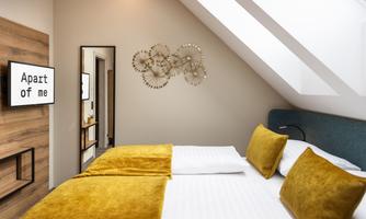 The image shows a modern bedroom with a cozy double bed and yellow pillows. A mirror hangs on the wall along with decorative wall art.