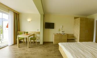 A bright and modern hotel room with a double bed, a table, and chairs. Large windows provide plenty of daylight and a view outside.