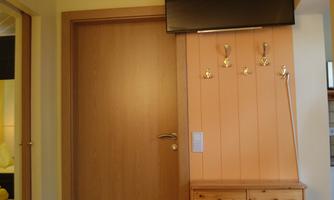 A cozy room with a wooden door, a television on the wall, and a small shelf. The wall is painted in warm shades of yellow.