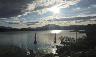 A quiet lake at sunset, surrounded by mountains. Some loungers and boats can be seen at the shore.
