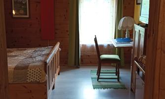 View through a doorway into a room with a wooden bed on the left, window with curtains, table with chair, bedside table with lamp, television and a red infrared heating panel on the wall; above the door hangs the sign 7. Himmel.
