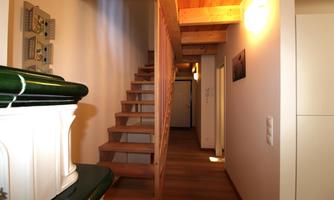 A bright hallway with a wooden staircase and warm light. The walls are plain, and the floor is made of wood.