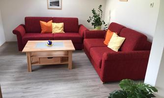 A cozy living room with red sofas and colorful cushions. A wooden table and a plant complement the friendly atmosphere.