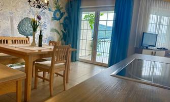 A light-filled living room with a wooden dining table and a large window. The walls are decorated with floral patterns and offer a view of the surroundings.