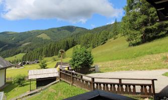 A picturesque mountain landscape with green meadows and wooded hills. In the foreground, several wooden houses and a path can be seen.