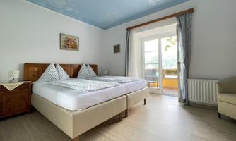 A bright bedroom with two beds and a window leading to a balcony. The walls are painted in soft colors, and the floor is made of light wood.
