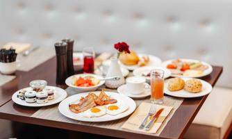 A hearty breakfast with fried eggs, bacon, and fresh fruit. Along with various jams, rolls, and drinks on the table.