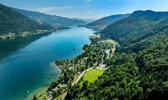 A beautiful lake surrounded by green mountains and forests. In the foreground are some houses and a paved path.