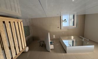A modern bathroom view with gray tiles and a small sink. The window provides natural light and a green mat is on the floor.