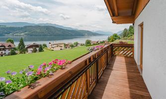 A balcony with a wooden railing and colorful flowers. In the background, there is a lake and green hills.