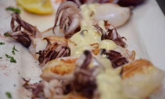 A delicious plate of grilled octopus, served with a creamy sauce. Next to it are lemon slices and fresh vegetables.