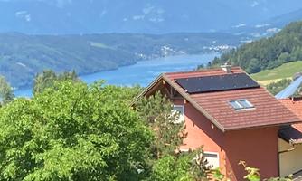 A picturesque house with solar panels on the roof, surrounded by lush greenery. In the background, a beautiful lake and mountains stretch out.