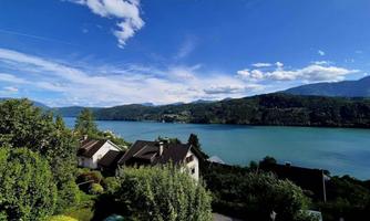 A picturesque landscape with a clear blue lake and green hills. In the foreground, there are some houses and lush plants.
