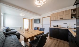 A modern kitchen with a wooden dining area and black chairs. The walls are bright and the room has large windows for plenty of natural light.