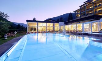 A modern hotel with a illuminated outdoor pool. In the background, gentle hills and the evening sky are visible.