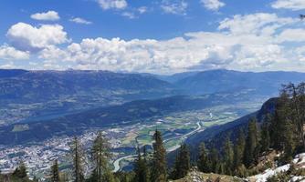 An impressive mountainous landscape overlooking a vast valley and a blue sky. Green meadows and forests surround the town in the valley.