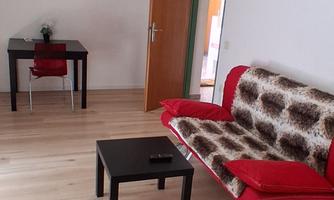 A simple room with a red sofa and a small table. In the background, a dining area with a table and a chair is visible.